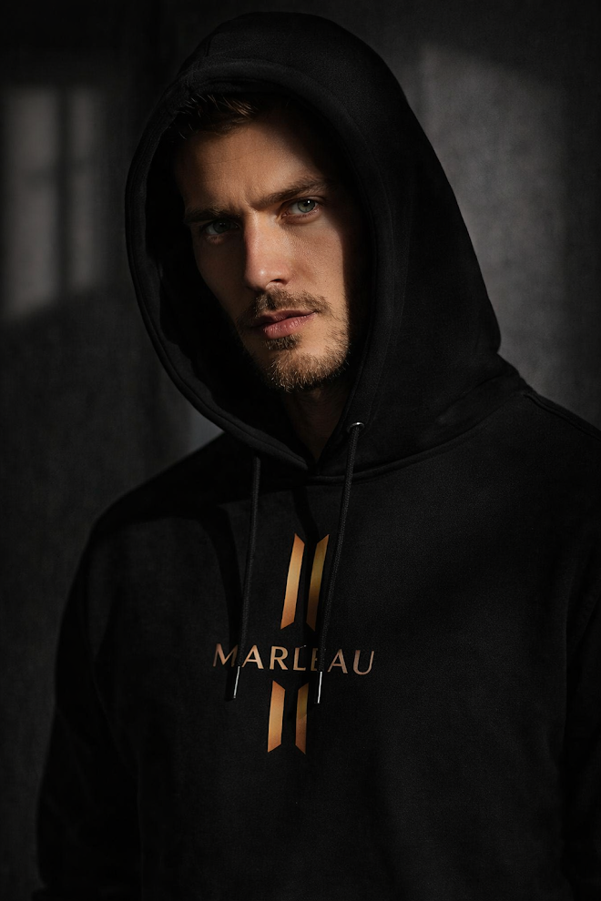 Male model wearing MARLEAU Axis Signature black hoodie with bronze front logo and hood up.